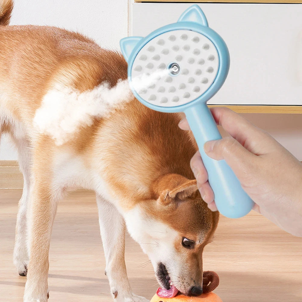 Hand holding a blue cat-eared pet grooming brush with mist feature, while a dog plays with a toy on the floor.